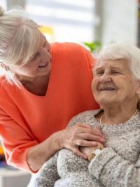 A caregiver with her arms around an older woman, both smiling.