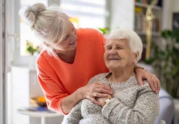 A caregiver with her arms around an older woman, both smiling.